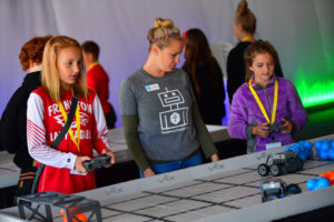 Two young girls control small robots on a VEX Robotics competition field, guided by an instructor wearing a “TechPoint Youth” shirt. The girls wear bright yellow lanyards, and the scene is set in a well-lit room with other participants in the background.