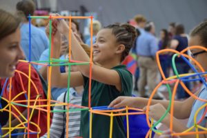 A young girl with braided hair smiles while building a colorful structure with interlocking plastic sticks. Other children and adults are participating in similar activities in the background.