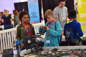 Two children, a boy and a girl, are actively engaged in building a structure using VEX Robotics parts. The boy, wearing a light blue hoodie, carefully places a piece, while the girl in a green jacket looks on with interest. Both are wearing red lanyards, and the background shows other participants and activity areas.