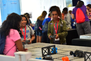 A group of children participate in a robotics event, operating robots on a VEX Robotics playfield. A girl in a green jacket and red lanyard, named Madison, controls a robot while another girl, wearing a blue t-shirt, watches and smiles. The background is filled with other participants and event activities.