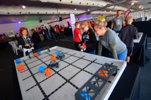 A group of young students participates in a robotics competition using remote-controlled robots on a VEX Robotics field. The field is marked with blue and orange objects. The students are focused and engaged, with one girl controlling a robot while others observe and participate. The event space is bustling with activity and other participants.