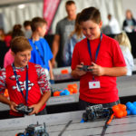 Two boys participate in a robotics competition, each holding a controller for their respective robots on a VEX Robotics playfield. Both boys are dressed in red shirts, with one wearing a patterned shirt and the other a solid color. They appear focused and engaged, with other participants and event staff visible in the background.