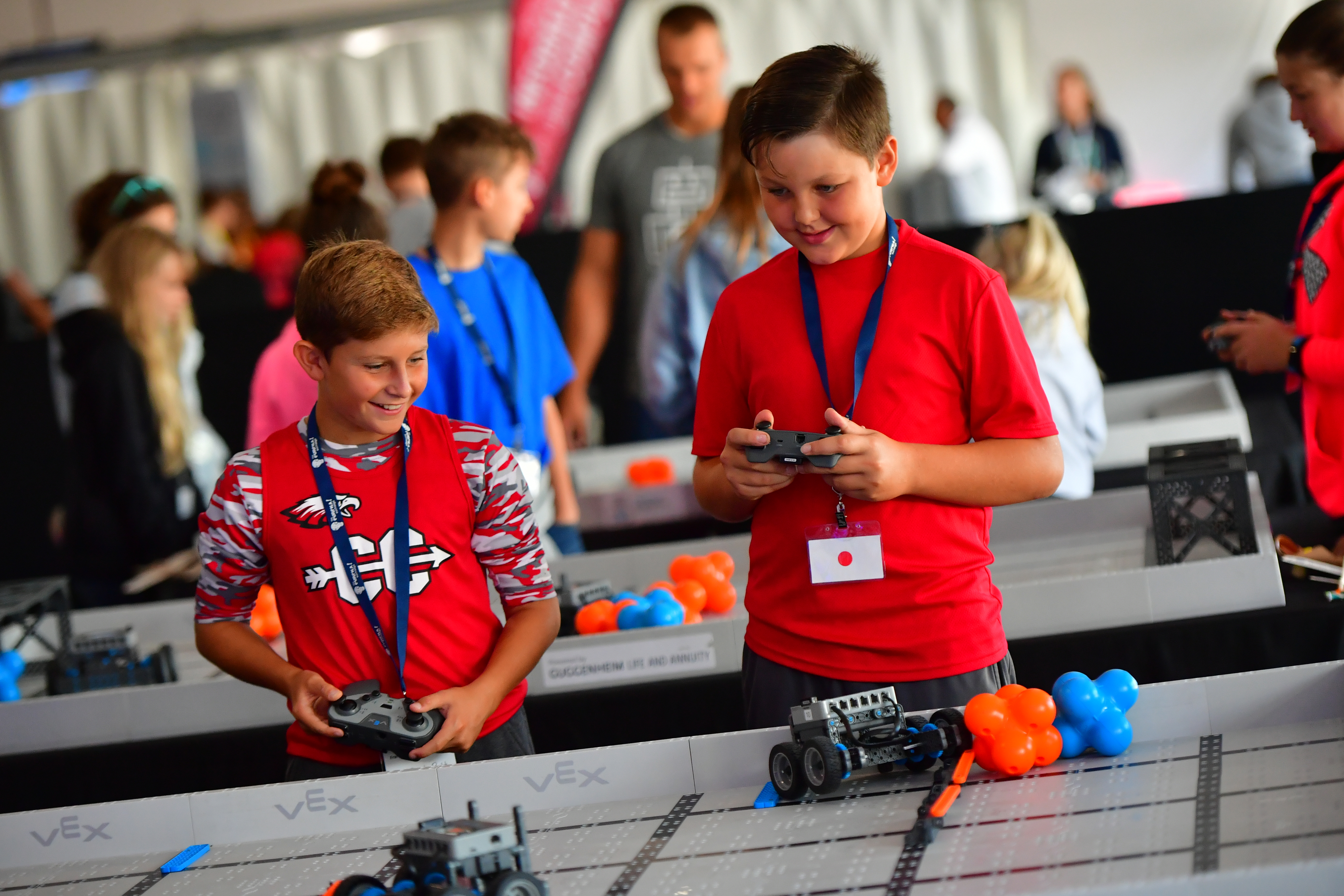 Two boys participate in a robotics competition, each holding a controller for their respective robots on a VEX Robotics playfield. Both boys are dressed in red shirts, with one wearing a patterned shirt and the other a solid color. They appear focused and engaged, with other participants and event staff visible in the background.