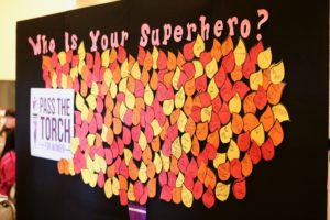 A wall display with the question “Who Is Your Superhero?” at the top, featuring numerous leaf-shaped notes in yellow, orange, and red. Each note has a name or message written on it, such as “My Mom” and “My Dad.” A “Pass the Torch for Women” sign is visible on the left side.