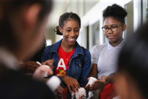 A group of young girls are engaged in a hands-on activity involving white, granular material, possibly snow or a science experiment. The girls are smiling and appear to be enjoying the experience. One girl is wearing a denim jacket over a red shirt, and another has her hair up in a bun and wears glasses.