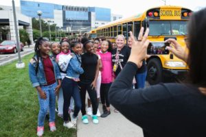 A group of smiling students poses for a photo in front of a yellow school bus. They are accompanied by a teacher or chaperone. The group stands on a sidewalk near a green lawn, and the background includes a modern building. Another person is taking the photo using a smartphone.