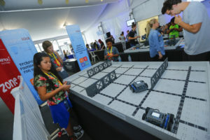 A group of children and adults participate in a robotics competition using remote-controlled robots on a VEX Robotics field. One girl in a floral shirt is focused on her robot, while others observe or engage in similar activities around the event space, which features banners and booths.