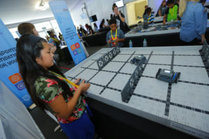 A girl with long black hair operates a remote-controlled robot on a large grid playfield at an event. Other children and adults are engaged in similar activities in the background, with banners and screens displaying event information.