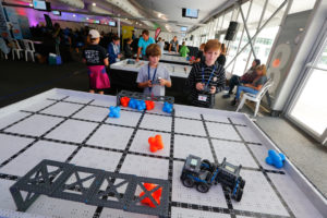 Two boys participate in a robotics competition, each controlling a robot on a VEX Robotics playfield filled with blue and orange objects. The boys are focused, one wearing a striped shirt and the other a gray t-shirt. The background shows other participants and spectators in a large, brightly lit room.