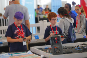 Two boys participate in a robotics workshop, examining parts and assembling components. One boy has purple hair and wears an Indianapolis 500 t-shirt, while the other has red hair and glasses, wearing a Notre Dame INTECH t-shirt. They both have red lanyards and are surrounded by various tools and materials.