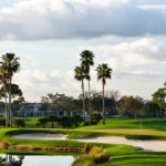 A banner featuring the Keiser University Golf Classic, with a logo depicting a golf ball and the text “Keiser University” above “Golf Classic.” The logo also includes a badge for “Kids in STEM.” The background showcases a lush golf course with palm trees, sand bunkers, and a cloudy sky.